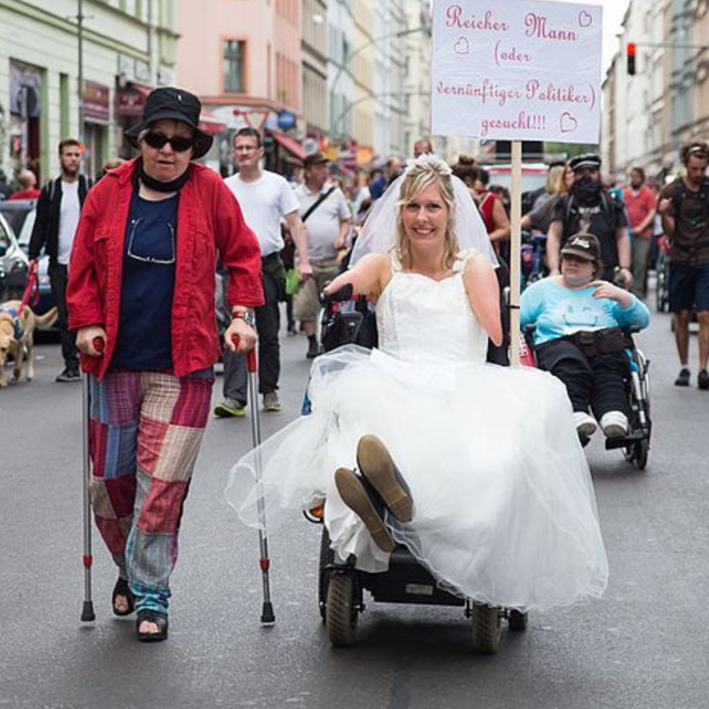 Short from a disability pride parade with a man wearing a top hat on crotches next to a woman in a wheelchair wearing a bridal dress.