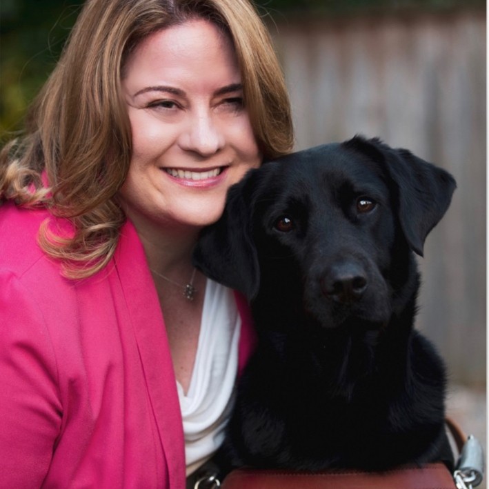 A smiling woman in a pink coat with brown hair and a guide dog. 