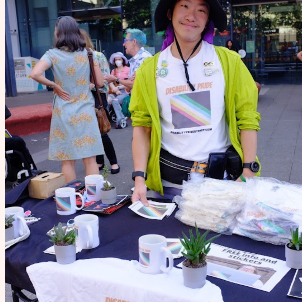 A man in a disability pride flag t-shirt sells disability pride flag merchandise 